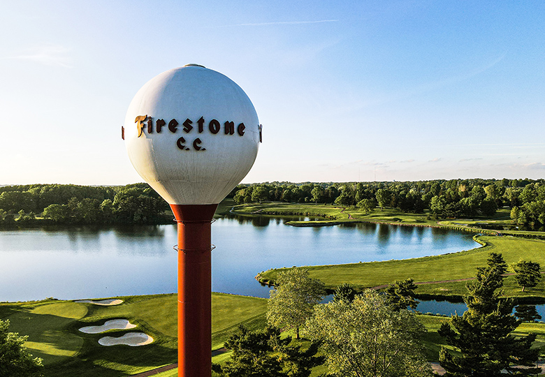 photo is of a golf course during sunset. At forefront of the image is a water tower in the shape of a golfball that says "Firestone C.C."