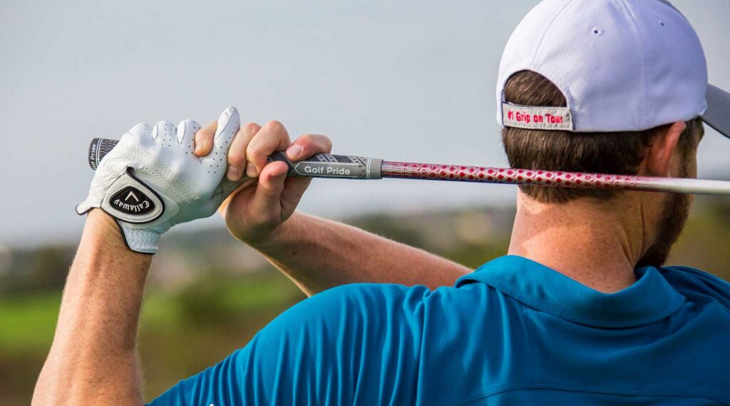 photo is of the back upper half of a male golfers body, at the end of his swing. visible are the words "Golf Pride" on the handle of his golf club.